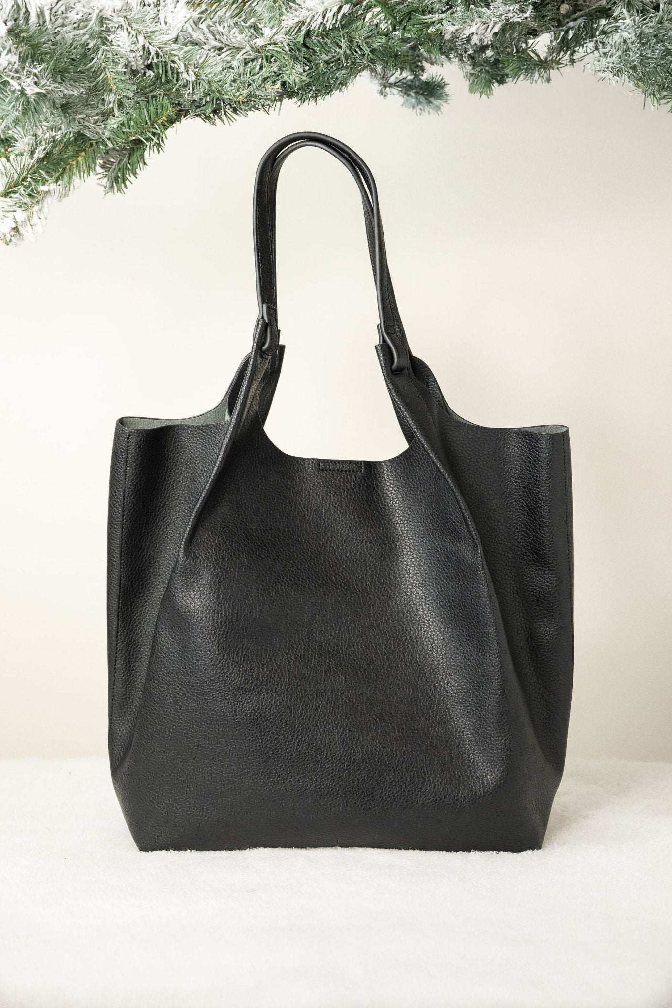 The Black Maeve Pebbled Leather Tote Bag rests on a white surface, accented by frosted evergreen branches above.