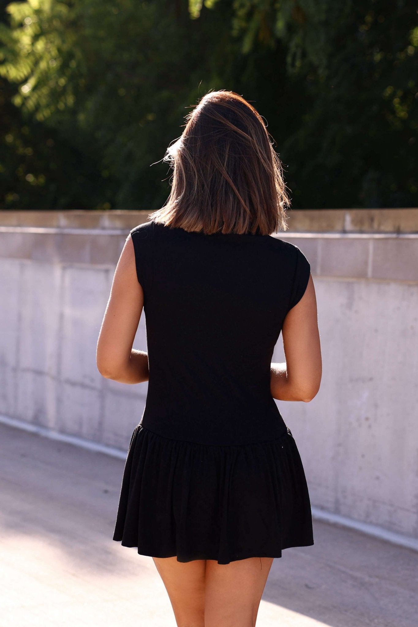 A woman in a Black Mock Neck Flare Mini Dress stands outdoors with her back to the camera on a sunny day.
