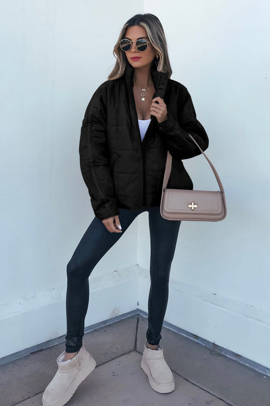 Woman in sunglasses, a Black Quilted Zip Up Puffer Jacket, leggings, and beige boots stands by a white wall on a concrete floor.