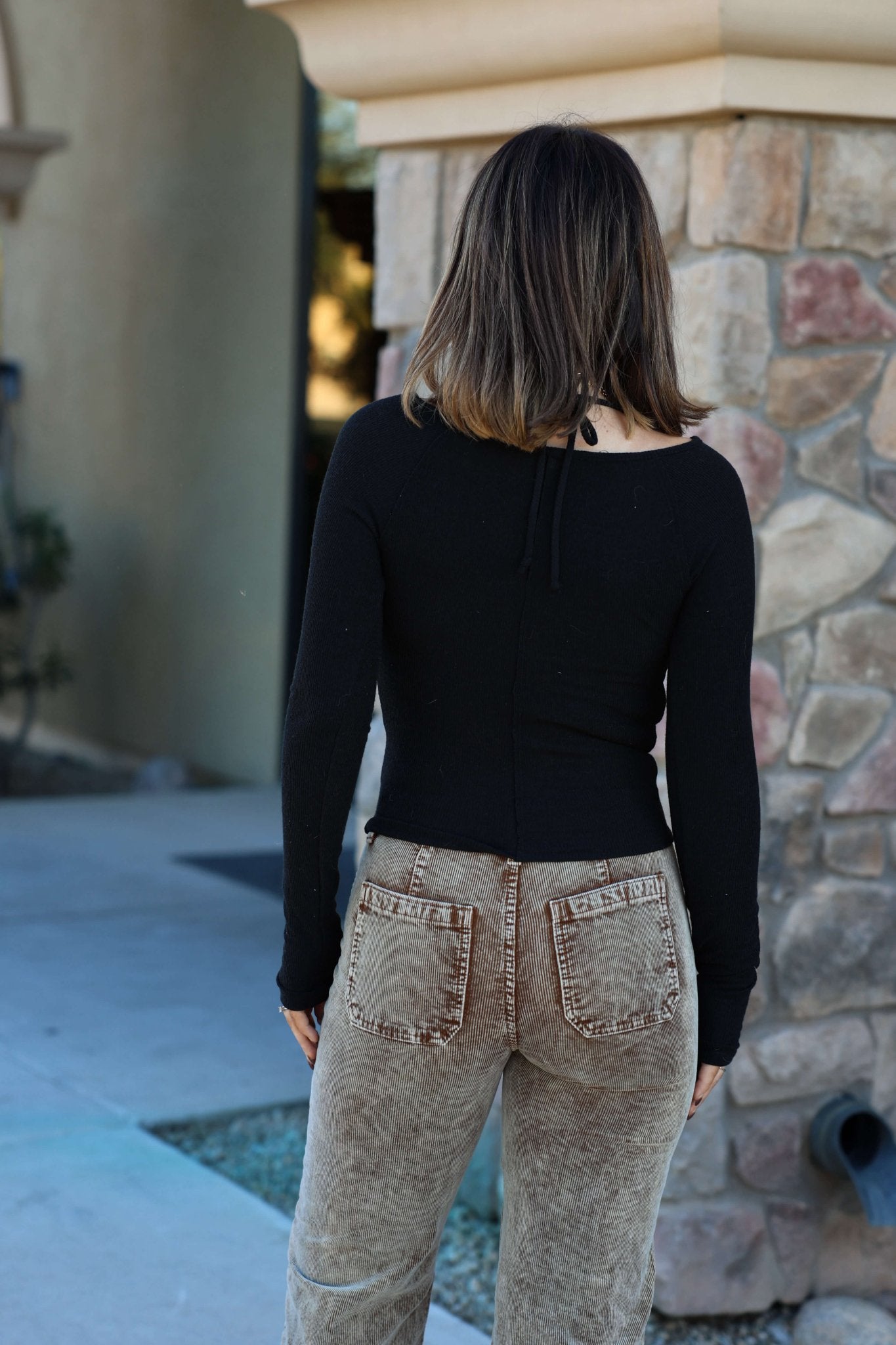 A woman with shoulder-length brown hair stands outdoors, facing away in a Black Ruched Long Sleeve Top.