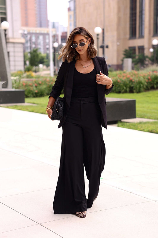 A woman in sunglasses wears the Black Ruched Sleeve Blazer with wide-leg pants and a top while walking outdoors on a sunny day.