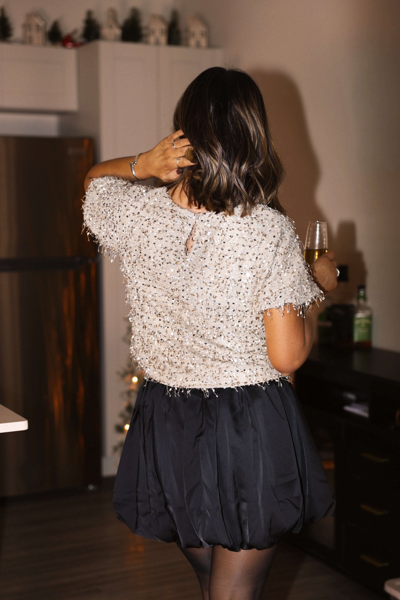 A woman wearing a Black Satin Pleated Mini Skirt holds a drink indoors, facing away from the camera in a sparkly top.