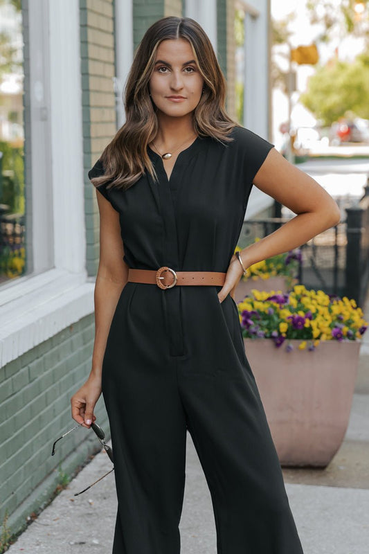 Confident woman in a Black Short Sleeve Belted Jumpsuit stands on the sidewalk, holding sunglasses with blooming flowers behind her.