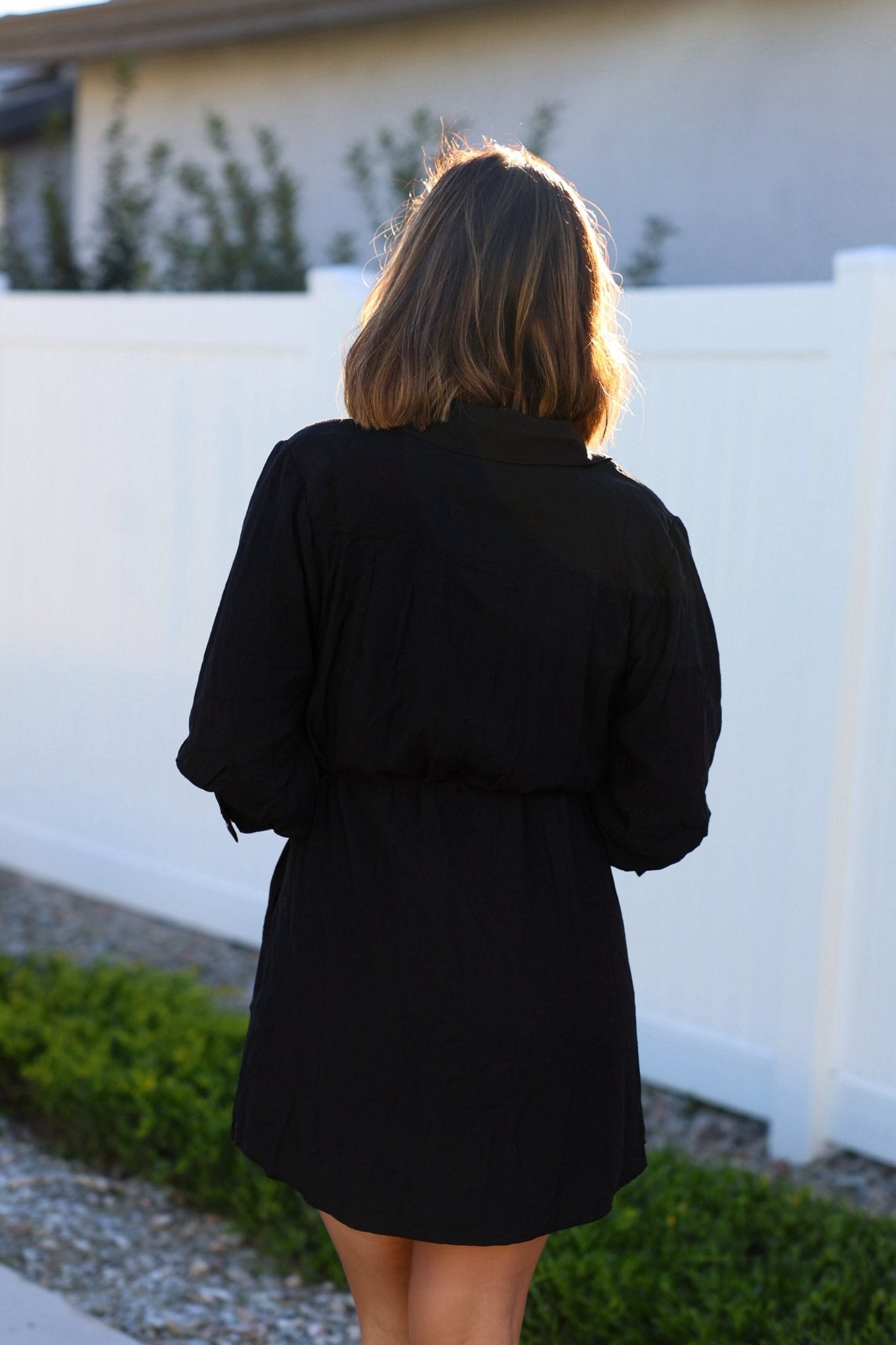 A woman with shoulder-length hair wears the Black Tie Mini Shirt Dress, standing outside by a white fence and green bushes.