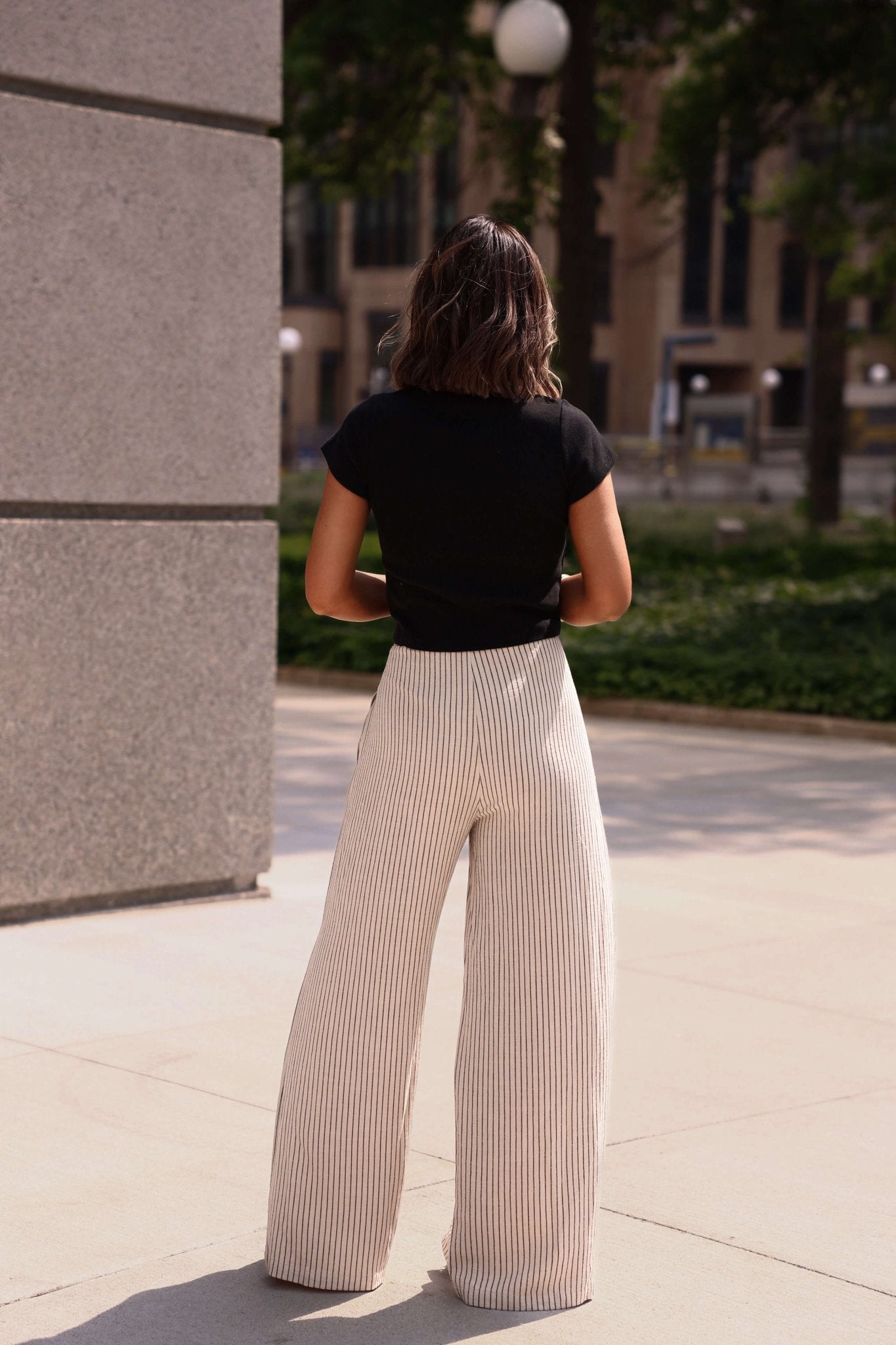 A woman with shoulder-length hair wears Blair Oatmeal Pinstripe Wide Leg Pants and a black top, standing outdoors and facing away.