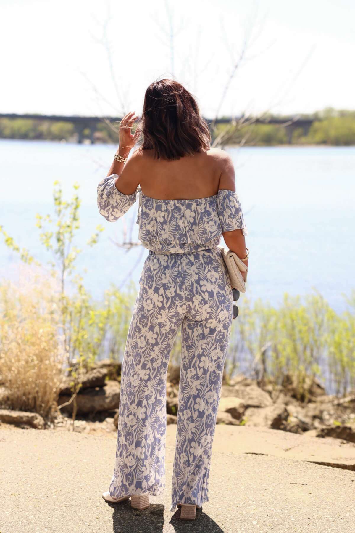 Woman in blue floral print linen pants (DOORBUSTER) stands by the water, facing away, holding a clutch purse.