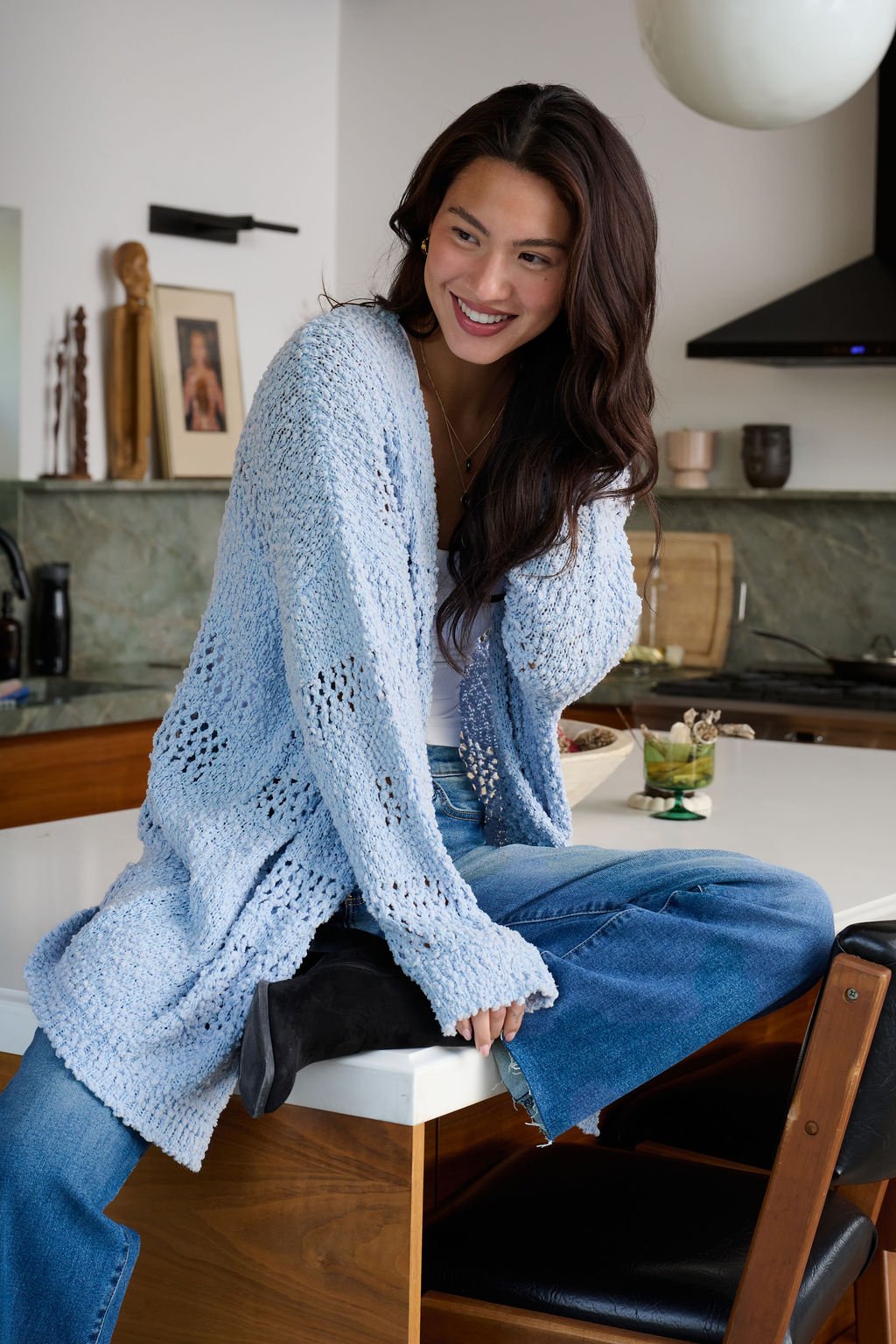 A woman in a Blue Open Front Popcorn Knit Cardigan and jeans sits smiling on a kitchen counter, legs crossed.