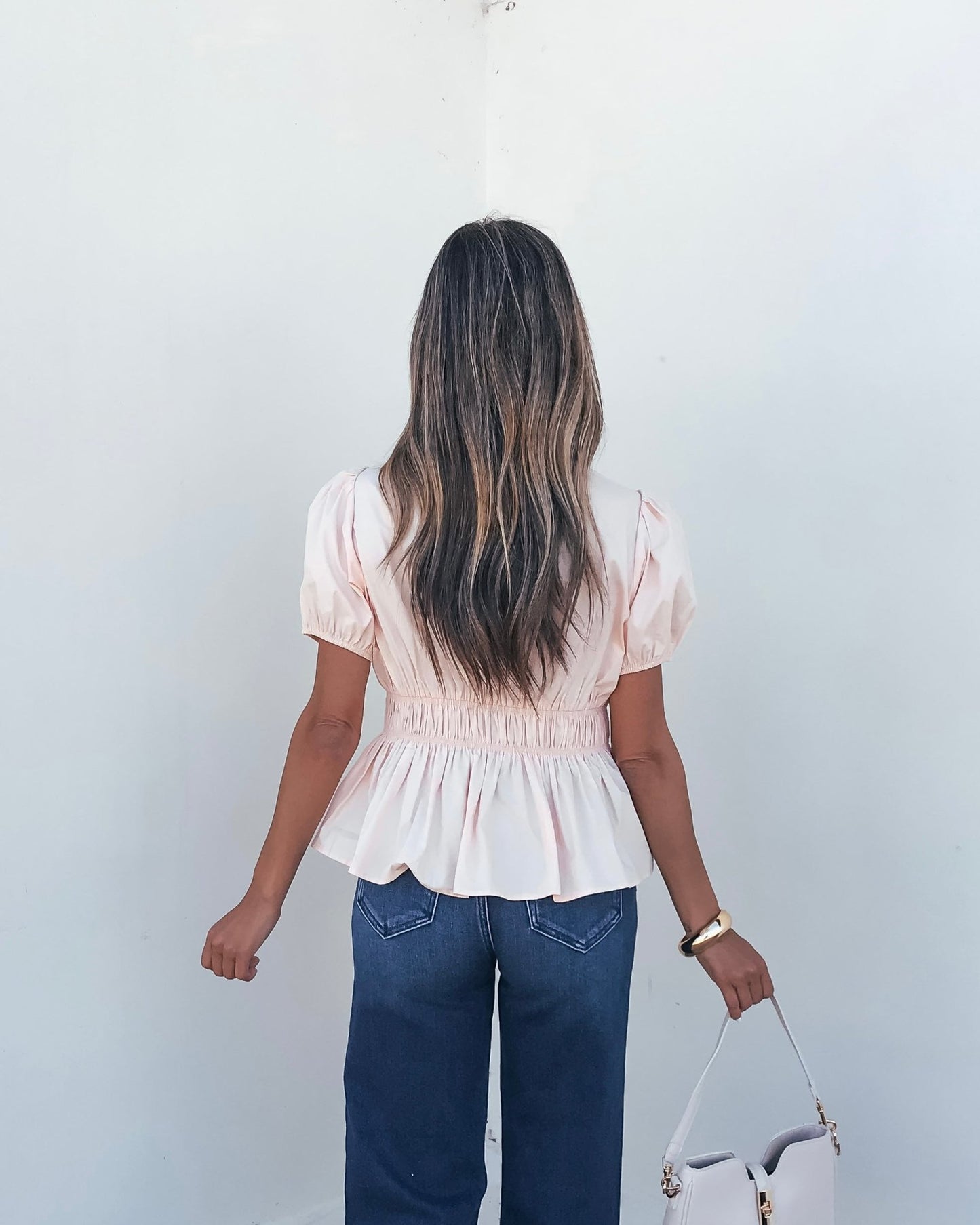 A woman with long wavy hair wears the Blush Puff Sleeve Peplum Top and blue jeans, holding a beige handbag by a white wall.