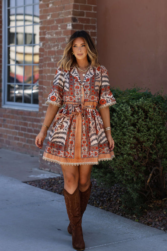 Smiling woman in a Botanical Rust Print Belted Mini Dress and brown boots stands by a brick wall on the sidewalk.