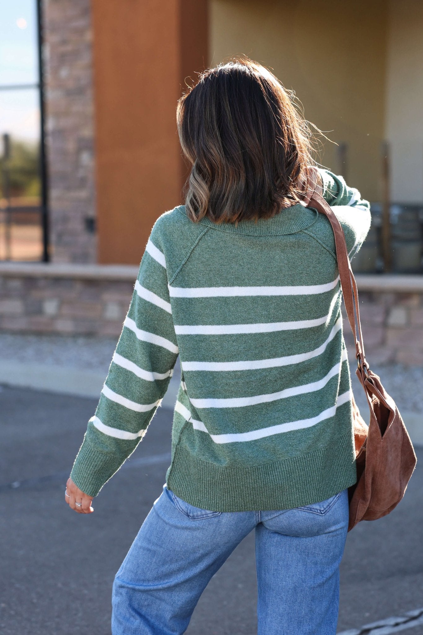 Wearing the Brinkley Sage and White Striped Sweater, someone walks outside with jeans and a brown shoulder bag for fall style.