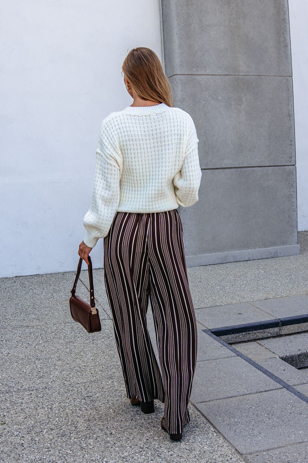 Woman with long hair in a white sweater and Brown & Cream Striped Drawstring Pants walks on the sidewalk carrying a brown handbag.
