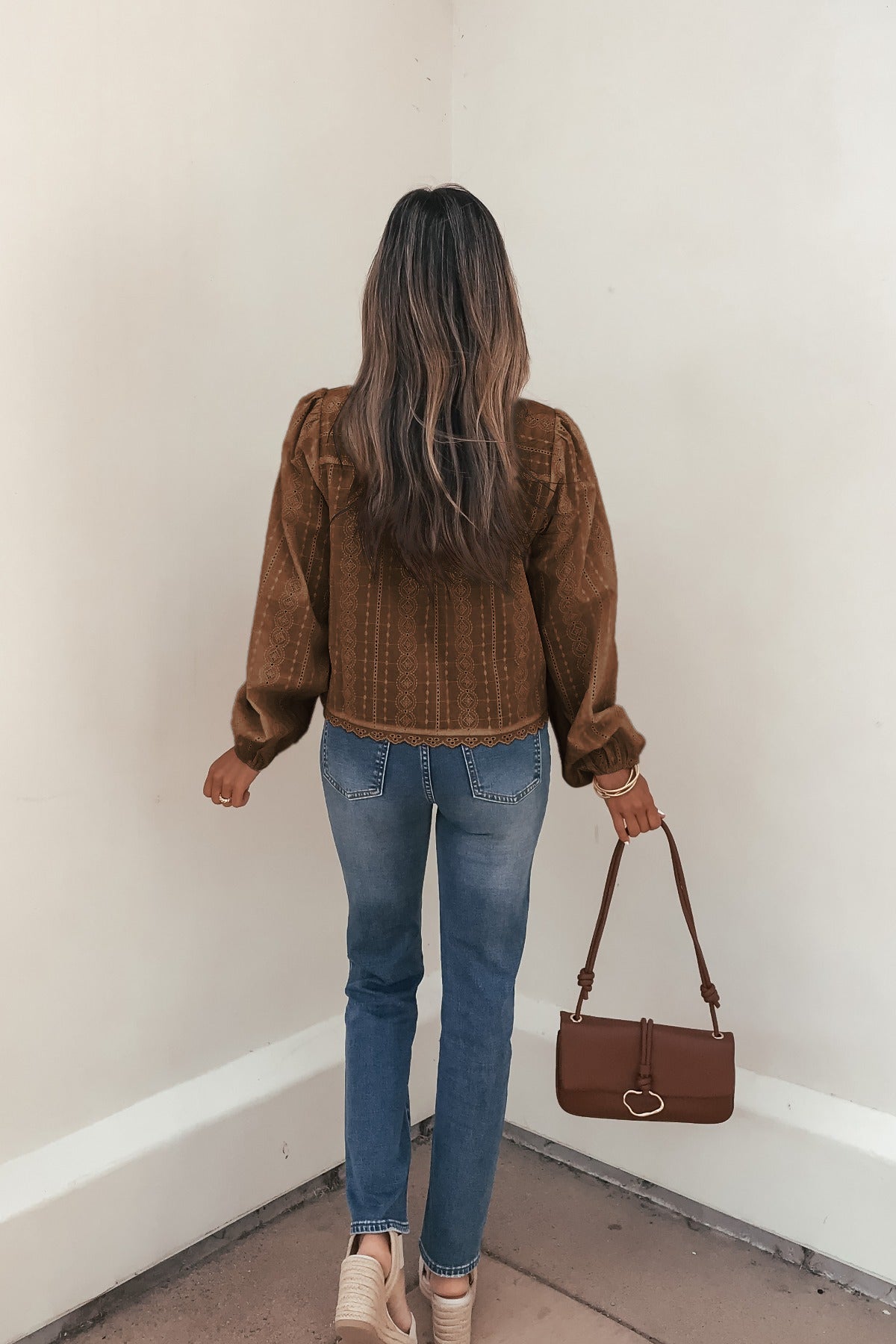 A woman with long hair in a brown eyelet tie front top, jeans, and sneakers stands facing a wall, holding a brown handbag.