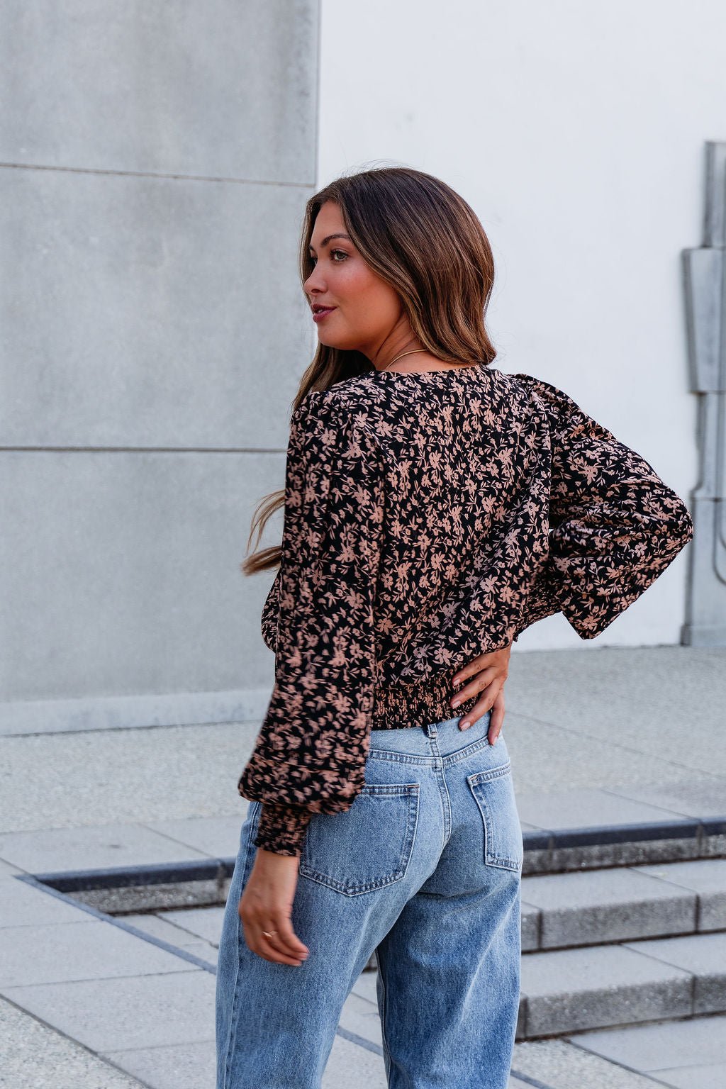 A woman wearing a Brown Floral Print Pleated Blouse and blue jeans stands outdoors, looking over her shoulder with confidence.