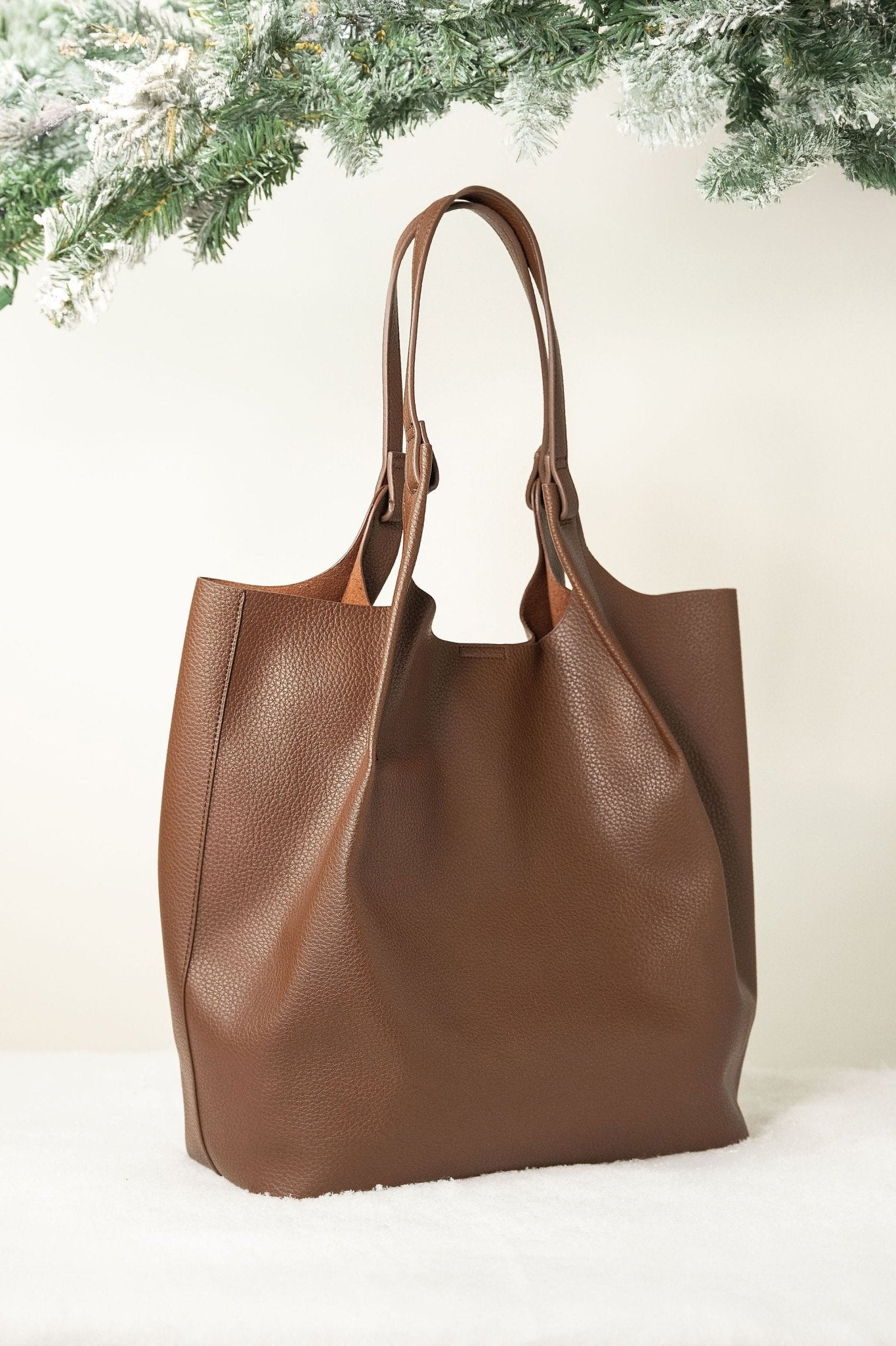 Brown Maeve Pebbled Leather Tote Bag displayed on a white surface, with green pine branches and snow behind.