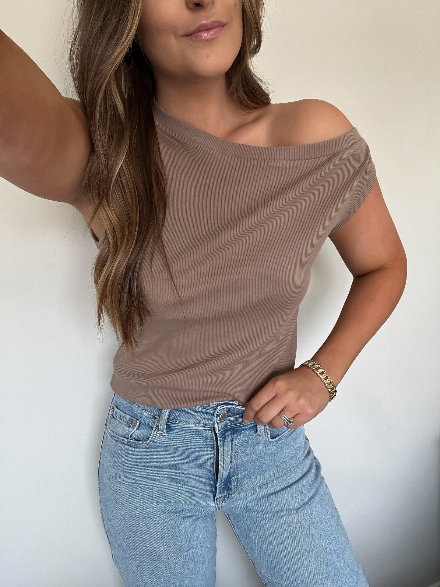 Woman in Brown Off The Shoulder Ribbed Top - FINAL SALE and light blue jeans, posing with one hand on her hip against a plain background.