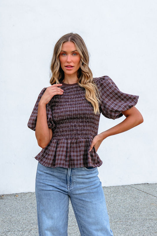 Woman wearing a Brown Puff Sleeve Peplum Top and jeans poses outdoors with one hand on her hip against a white wall.