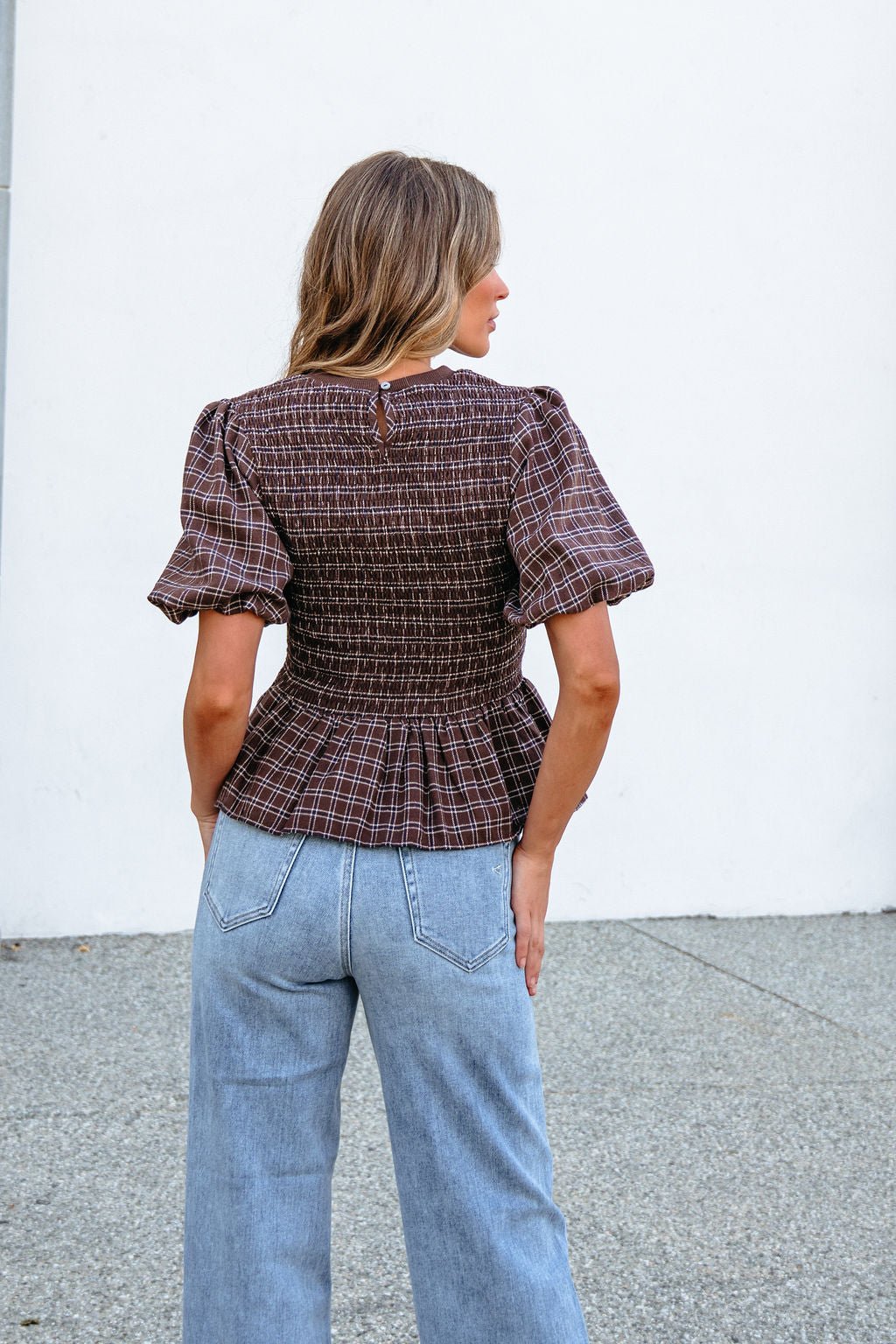 Woman with wavy hair, wearing a Brown Puff Sleeve Peplum Top and light blue jeans, stands on a textured surface, facing away.