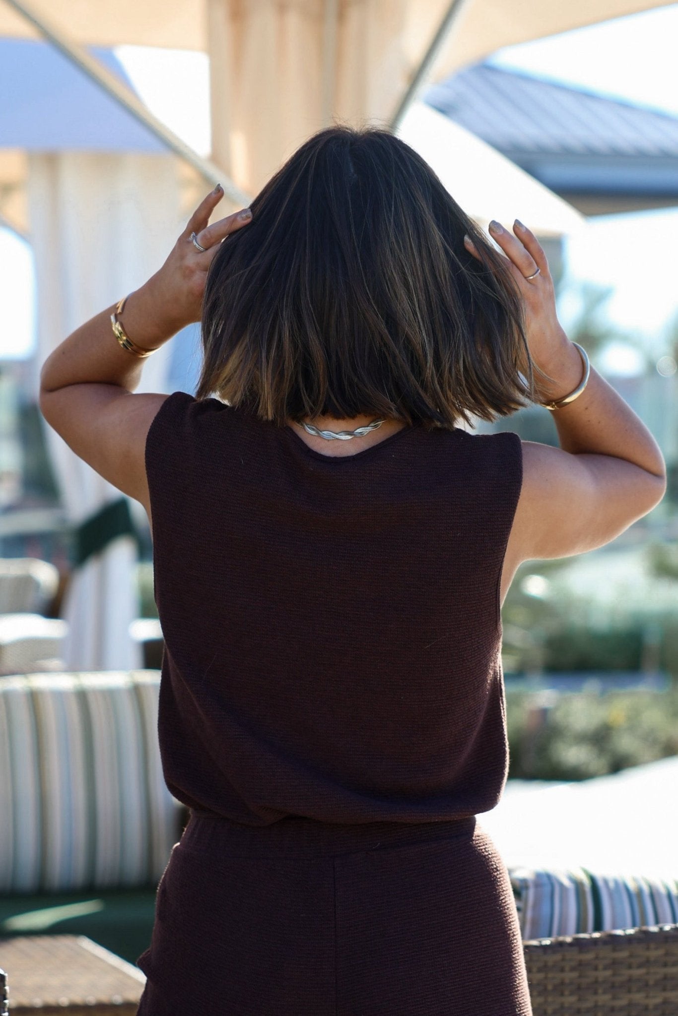 A person with shoulder-length dark hair touches their hair outdoors, wearing the Brown Sleeveless Bubble Hem Top.