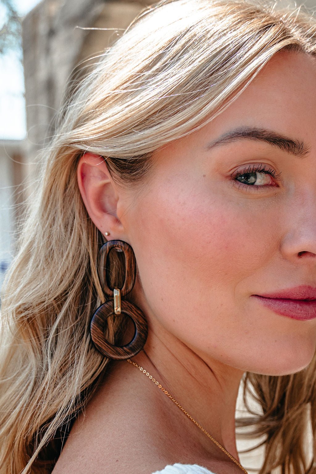 A woman with blonde hair models Brown Wooden Linked Dangle Earrings and a gold necklace in natural sunlight, facing the camera.