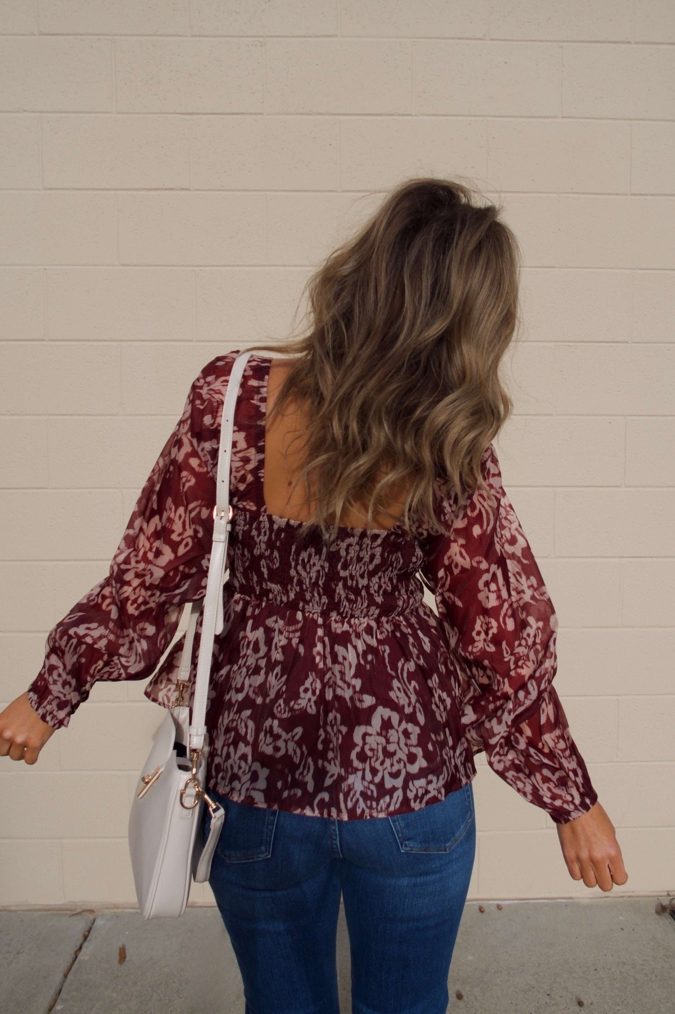Woman with wavy hair in a Burgundy Floral Chiffon Peplum Top, jeans, and white bag stands facing a beige wall, seen from behind.