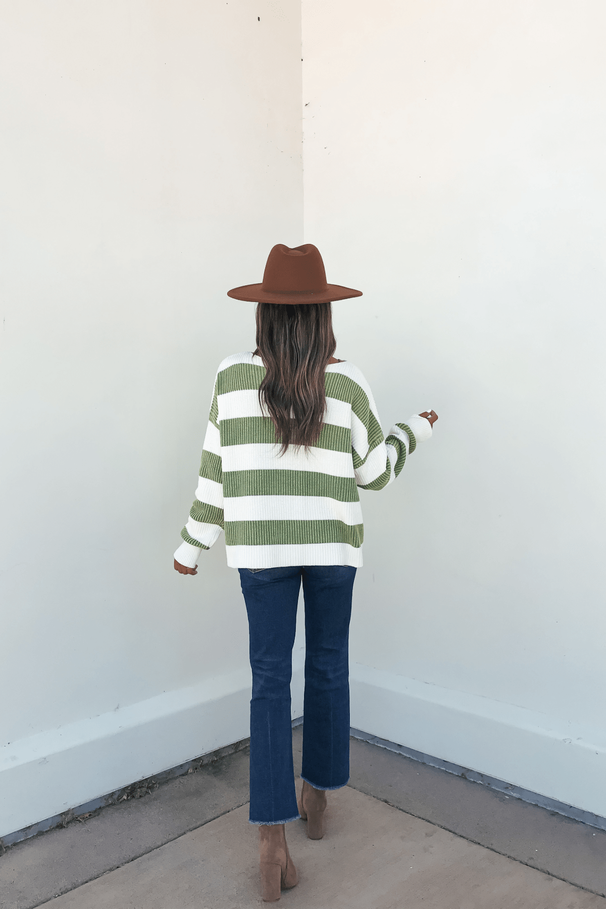 A person in a brown hat, Callahan Green and White Striped Sweater, jeans, and boots stands facing a white wall.