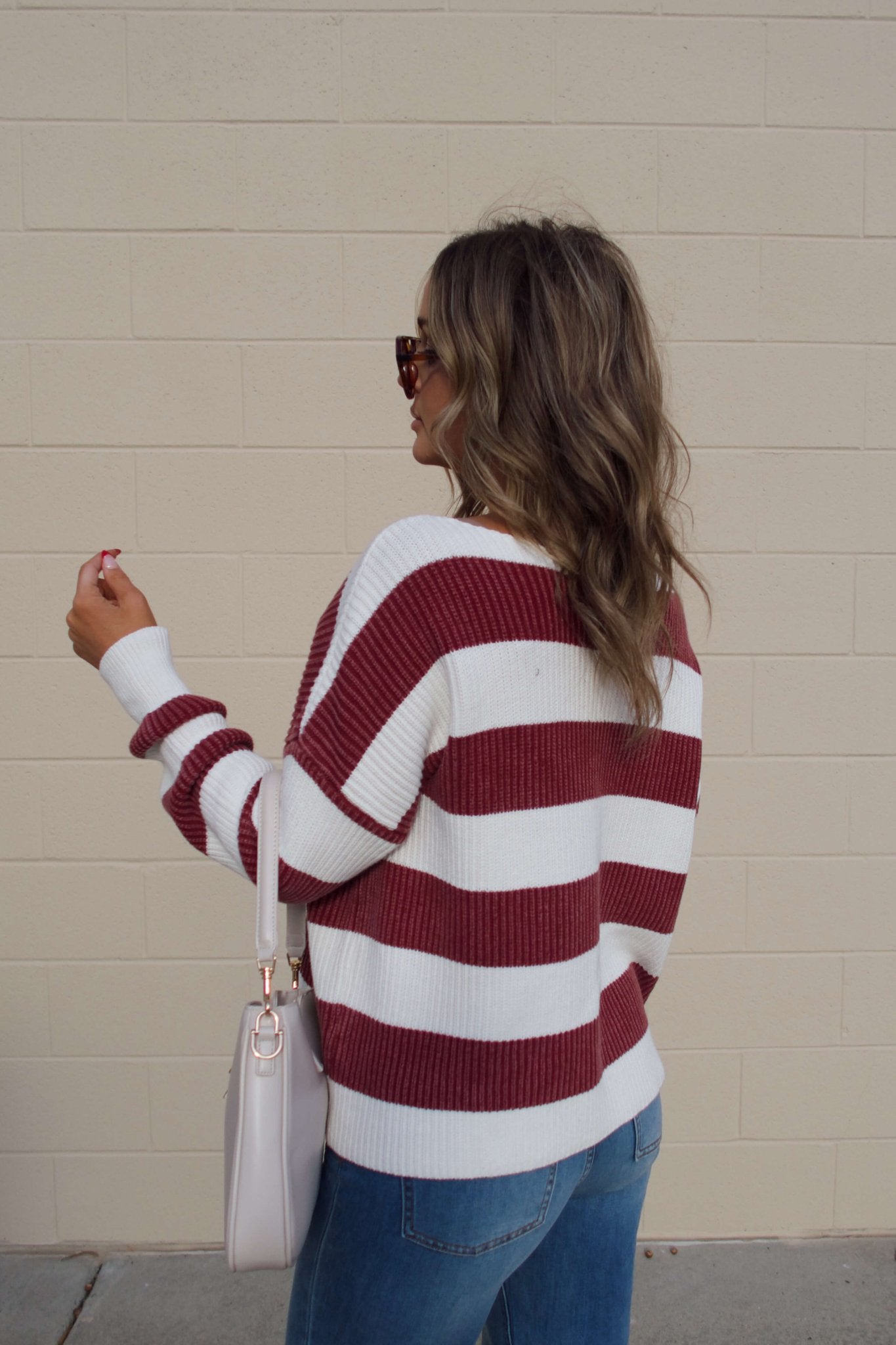 A woman in sunglasses wears a Callahan Wine and White Striped Sweater with jeans and a light pink handbag by a beige wall.