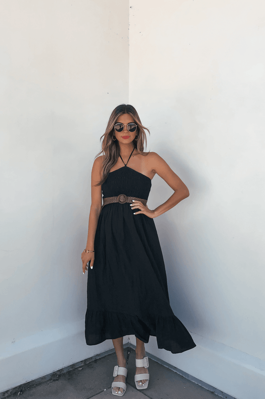 Woman wearing the Camille Black Halter Belted Midi Dress, white sandals, sunglasses, standing against a white wall.