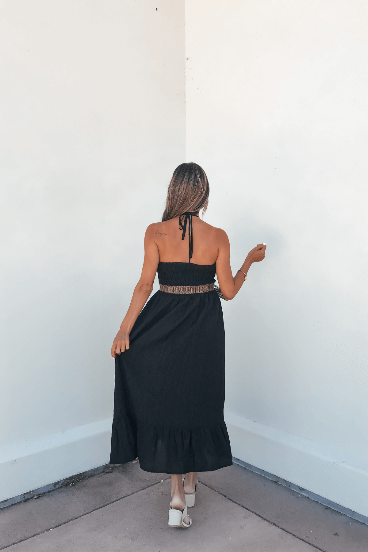 A woman wears the Camille Black Halter Belted Midi Dress, facing a white corner and holding her dress with one hand.