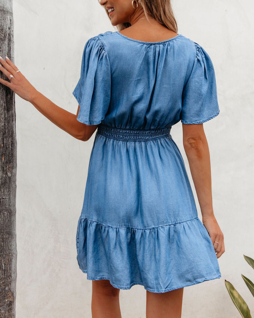 Woman in a Chambray Split Neck Smocked Mini Dress, standing sideways and touching a tree.