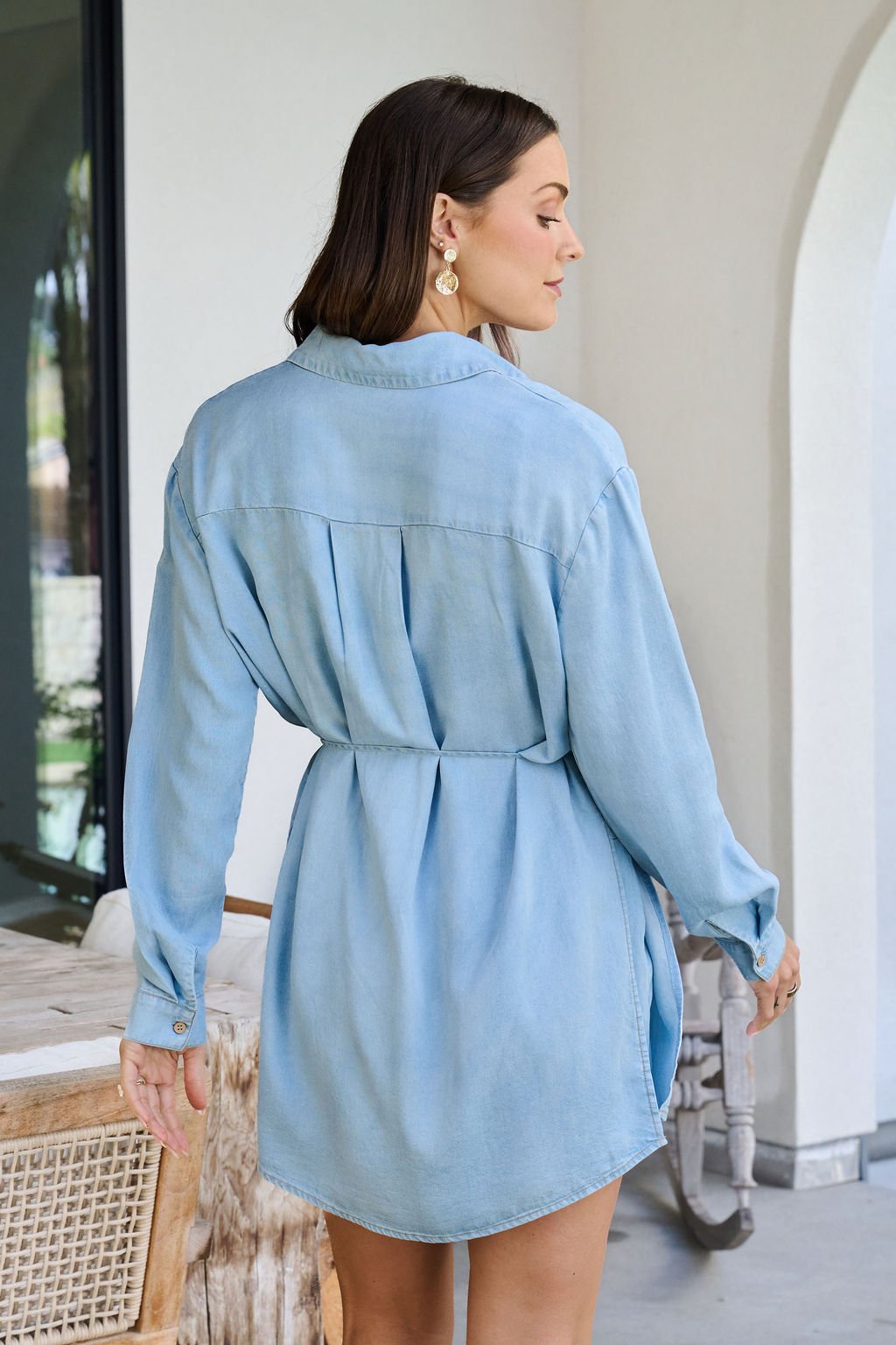 A woman in a light blue Chambray Tie Button Down Romper stands indoors, facing away and glancing over her shoulder.