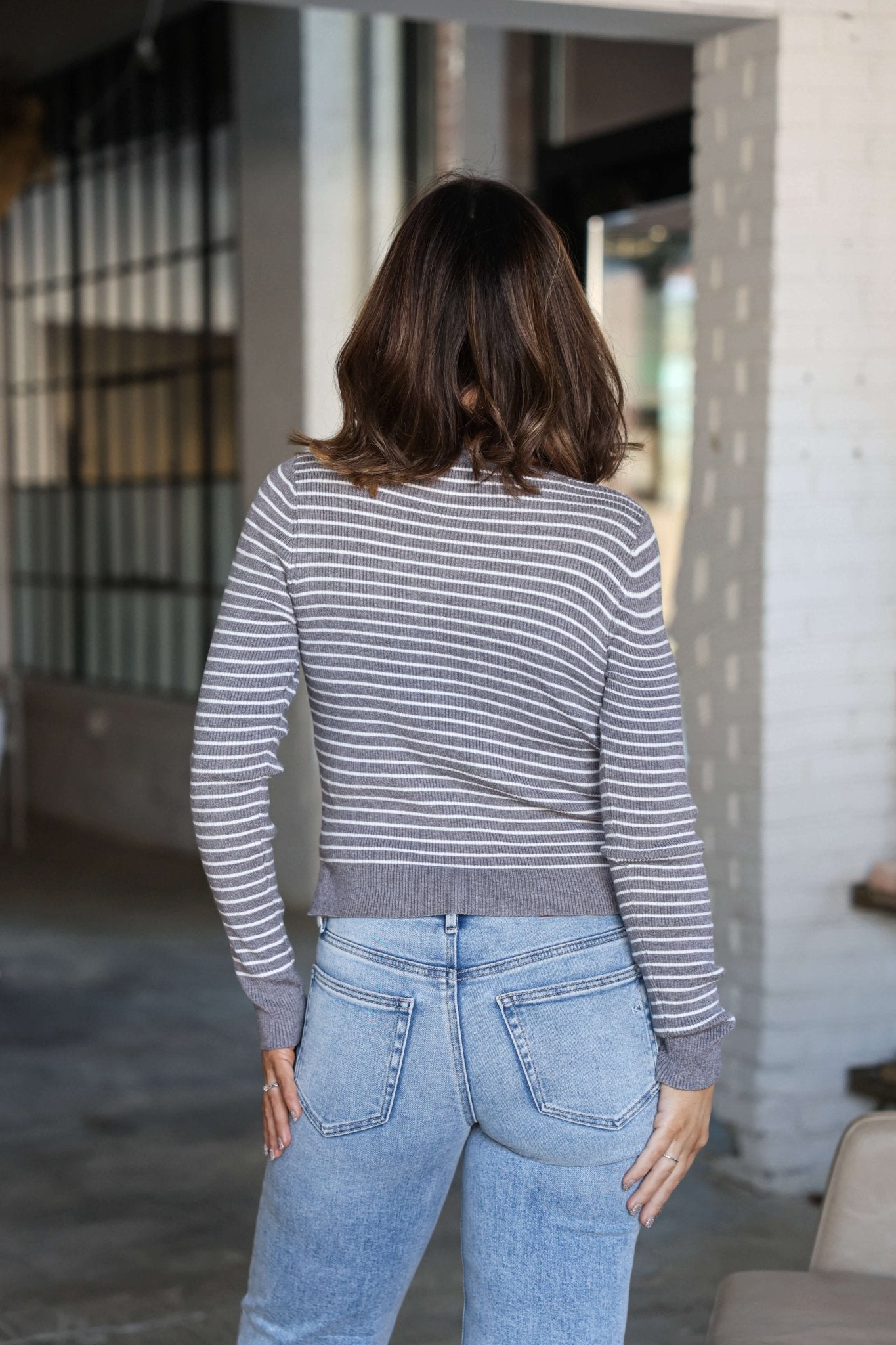 A woman with brown hair wears the Chelsea Grey and White Striped Sweater and blue jeans, standing indoors, facing away.