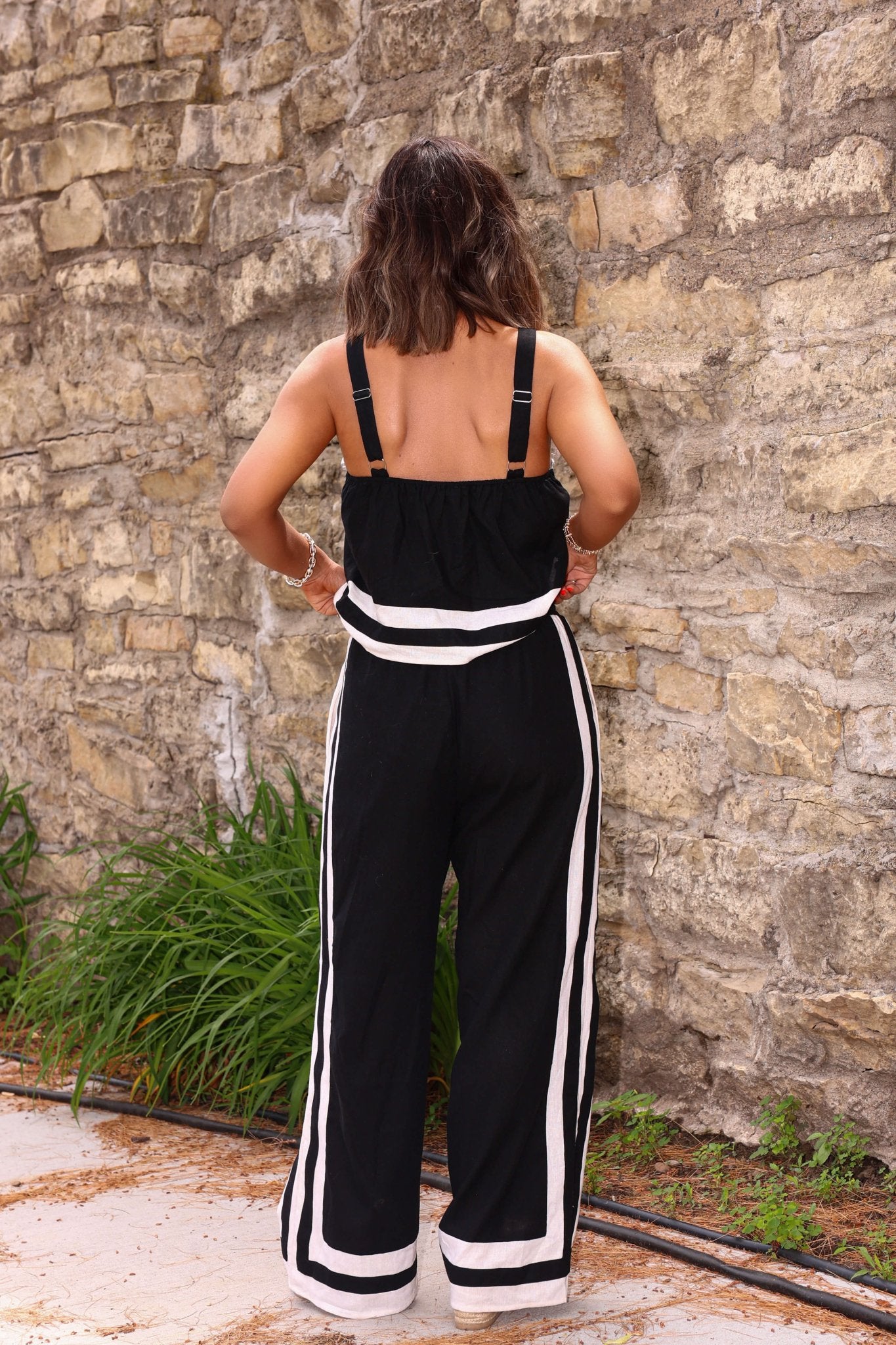 A woman stands with her back to the camera in Cheyanne Black and White Linen Pants, by a stone wall and greenery.