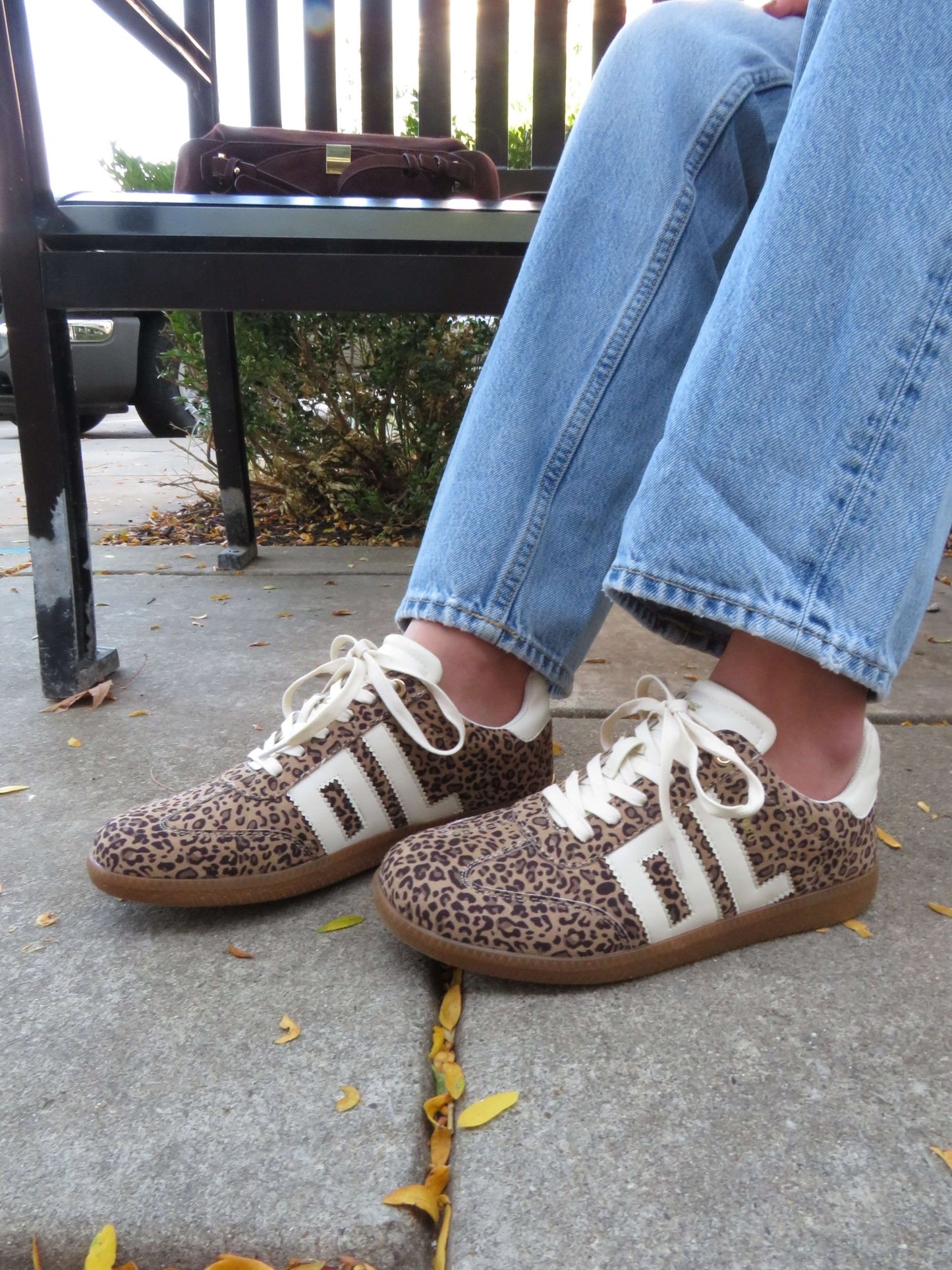 Wearing blue jeans and Chinese Laundry Trackstar Leopard Print Sneakers, sitting on a bench outdoors.