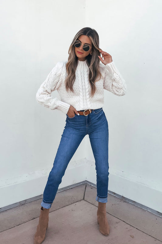 Woman in a Chunky Light Cream Mock Neck Sweater, blue jeans, and tan boots poses confidently against a plain white wall.
