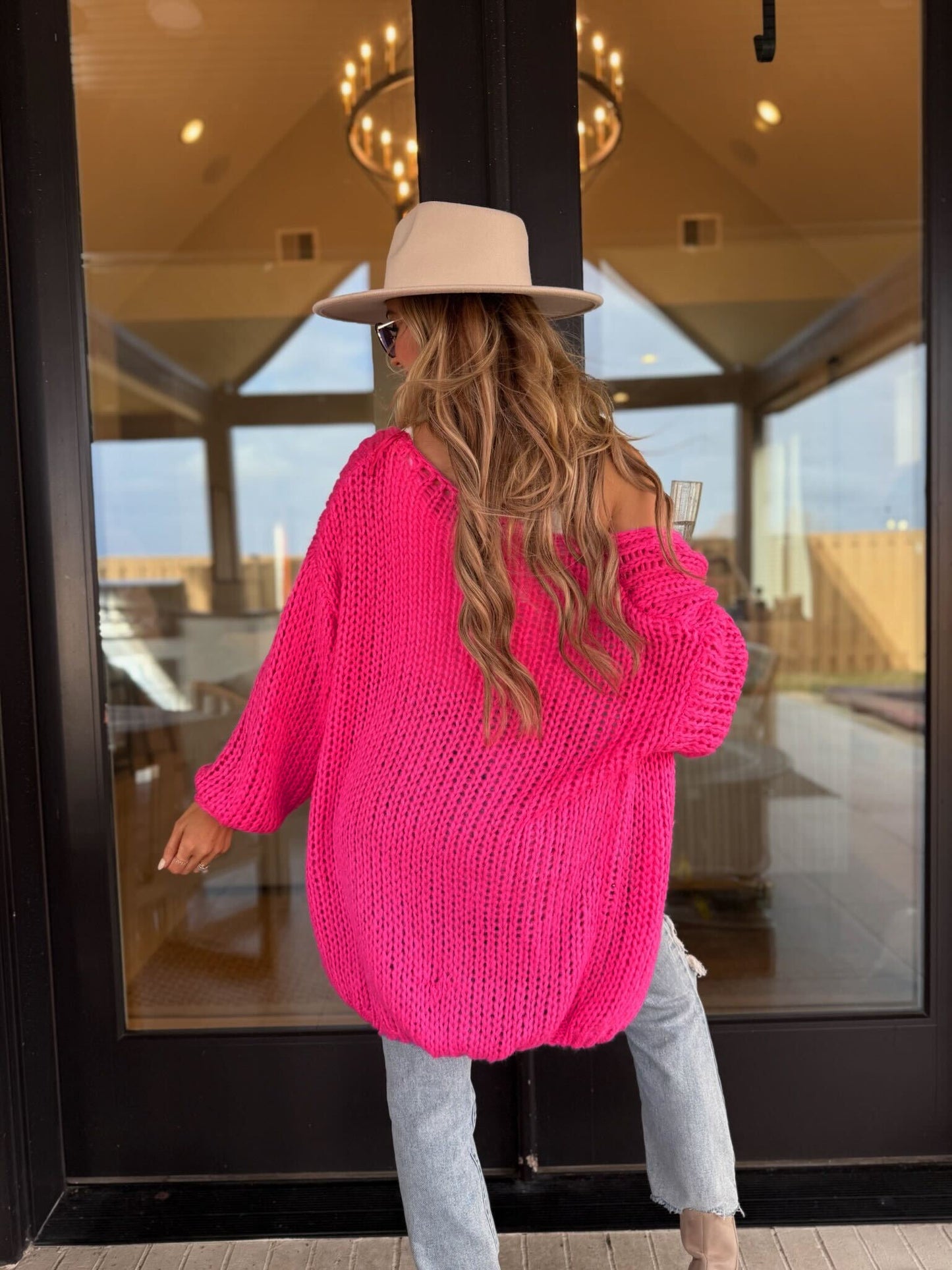 Woman with long wavy hair in a tan hat and ripped jeans wears a Chunky Pink Open Knit Sweater Cardigan, holding a drink by glass doors.
