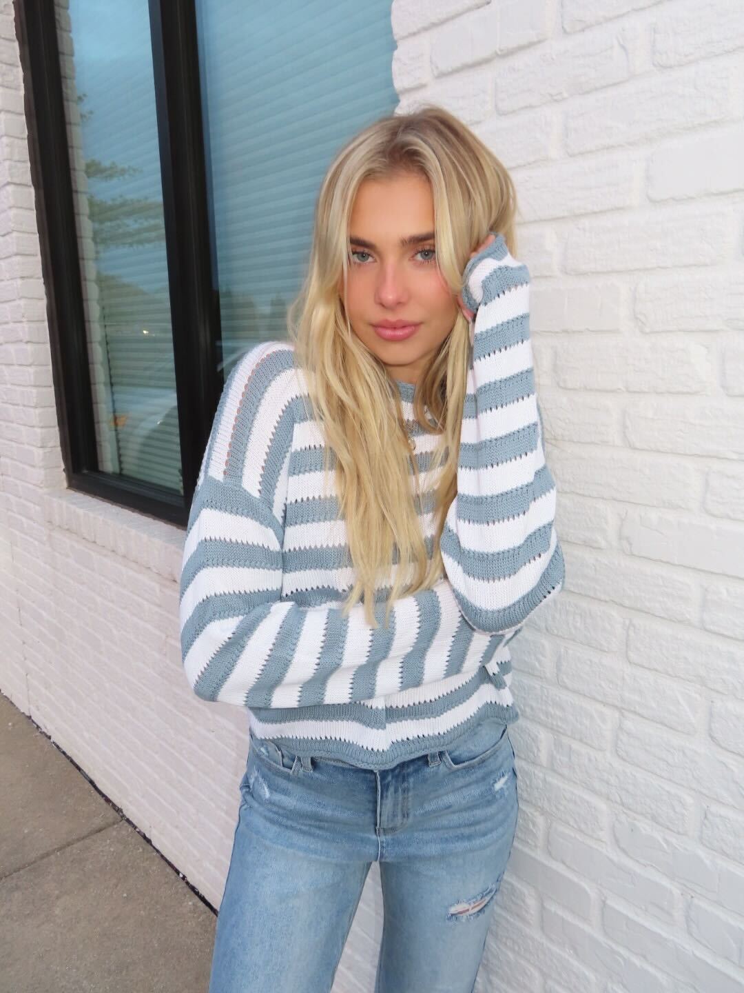 A woman wears the Cloud Grey and White Striped Sweater with jeans by a white brick wall, creating a timeless look.