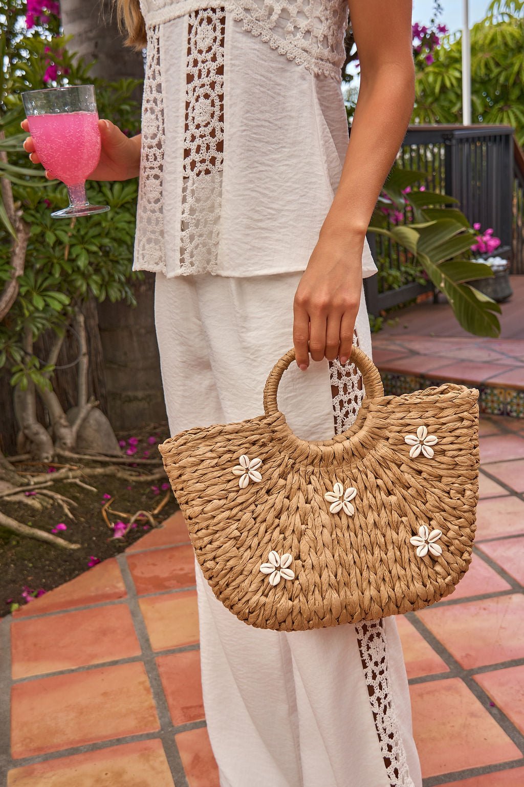 Woman in a white lace outfit holding the Coast Shell Tote - Natural with white flowers and a pink drink on a tiled patio.