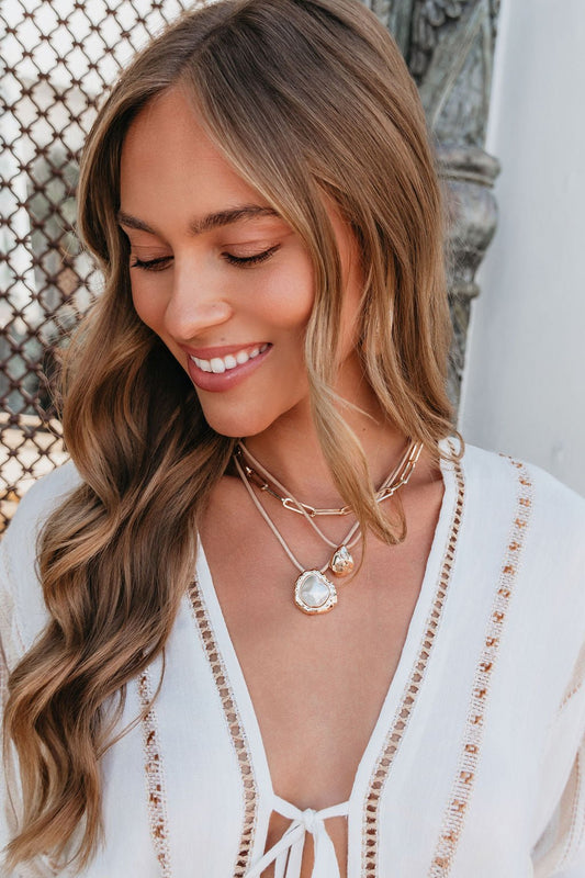 Woman with long wavy hair smiles in a white crochet top, wearing the Coastal Lux 3 Layered Necklace outdoors.