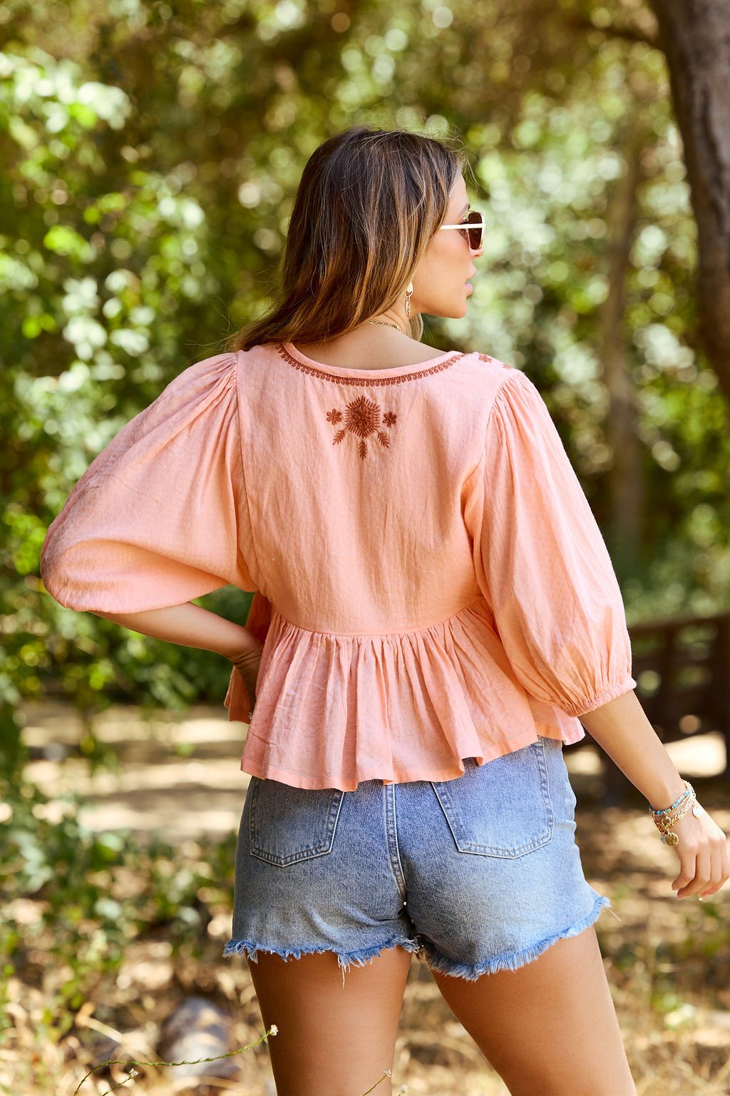 A woman outdoors in sunglasses and a Coral Tie Front Embroidered Peplum Top with puff sleeves and denim shorts, back to camera.
