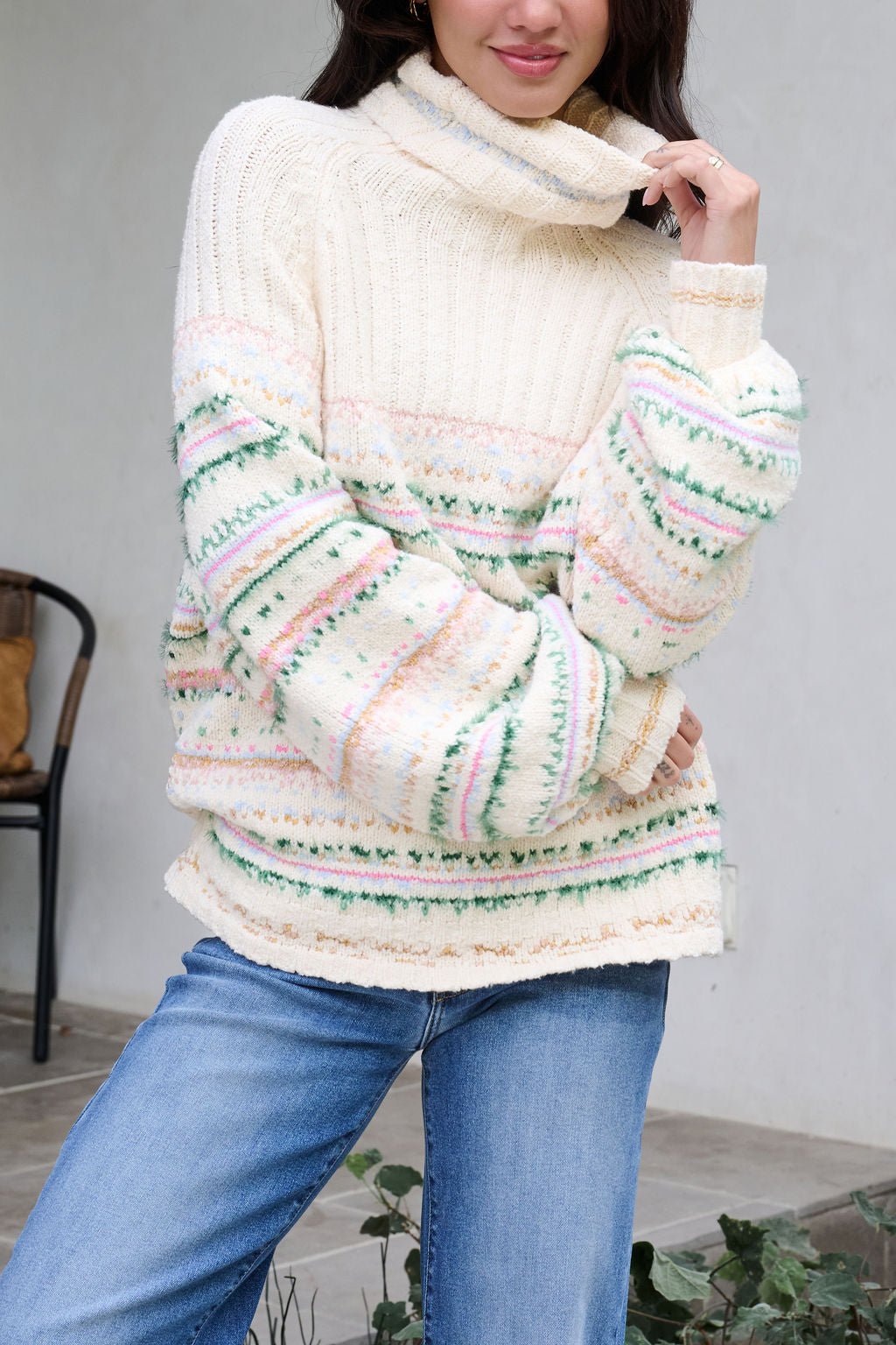 Woman in a Cozy Cream Turtleneck Striped Sweater with pastel stripes and blue jeans, radiating holiday spirit indoors by a chair.