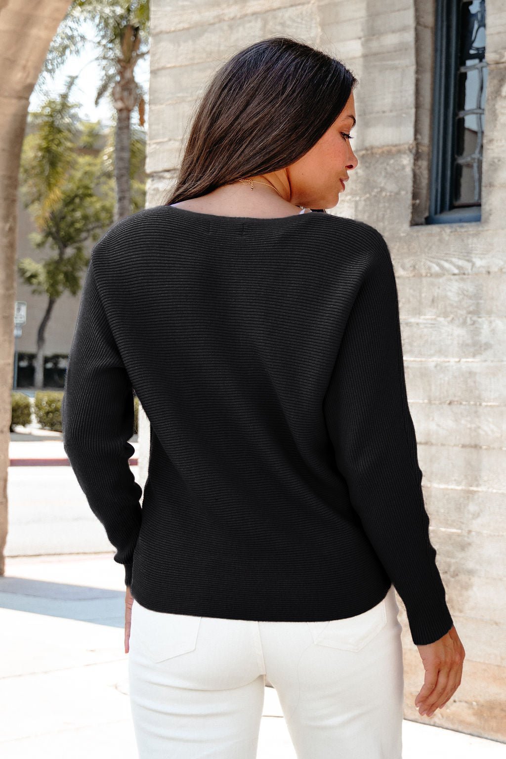 A woman with long brown hair wears the Cozy Ribbed Boat Neck Sweater - Black and white pants, standing by a stone building outdoors.