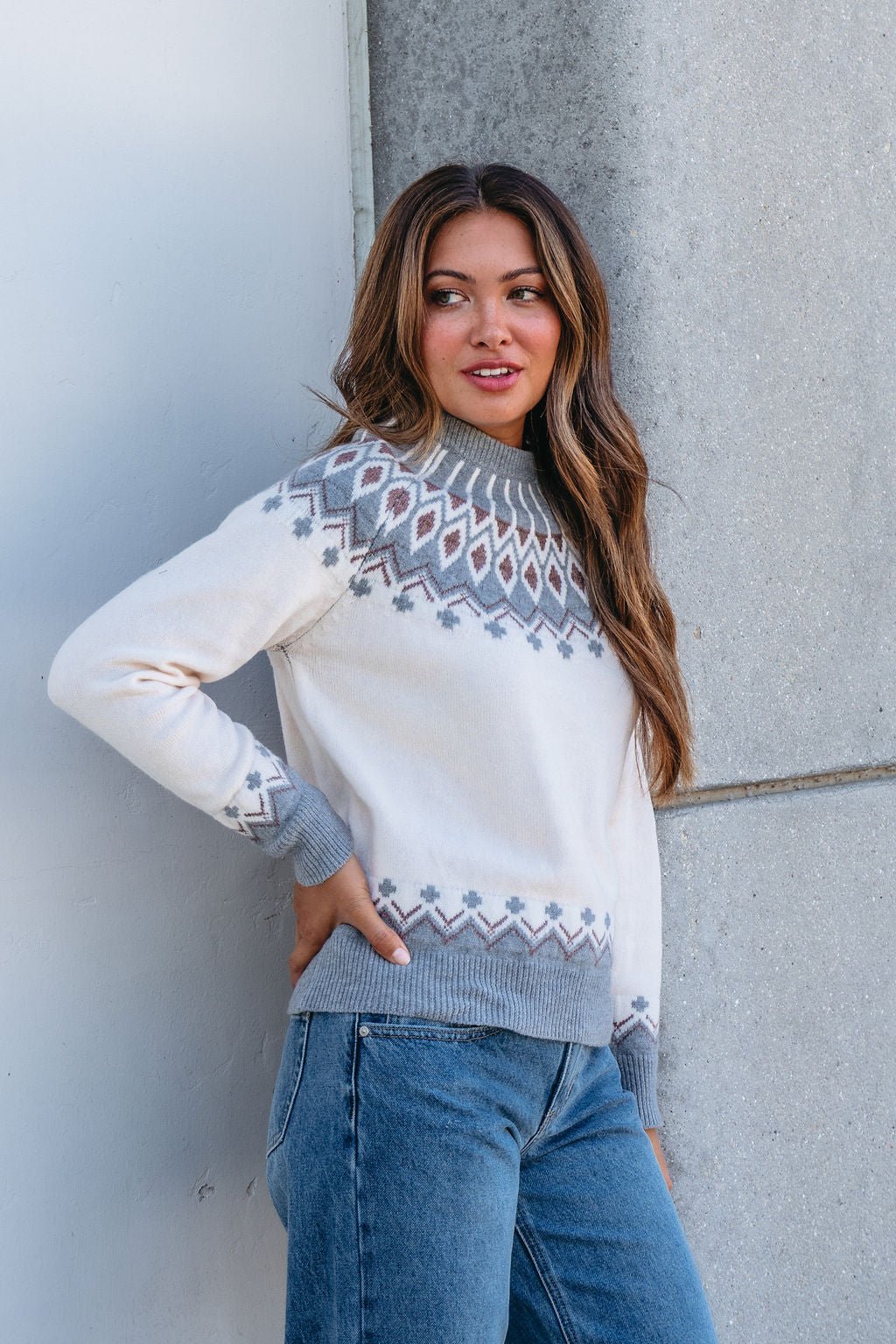 Woman in a Cream and Grey Aztec Print Sweater and blue jeans stands by a gray wall, smiling—ideal for autumn style.