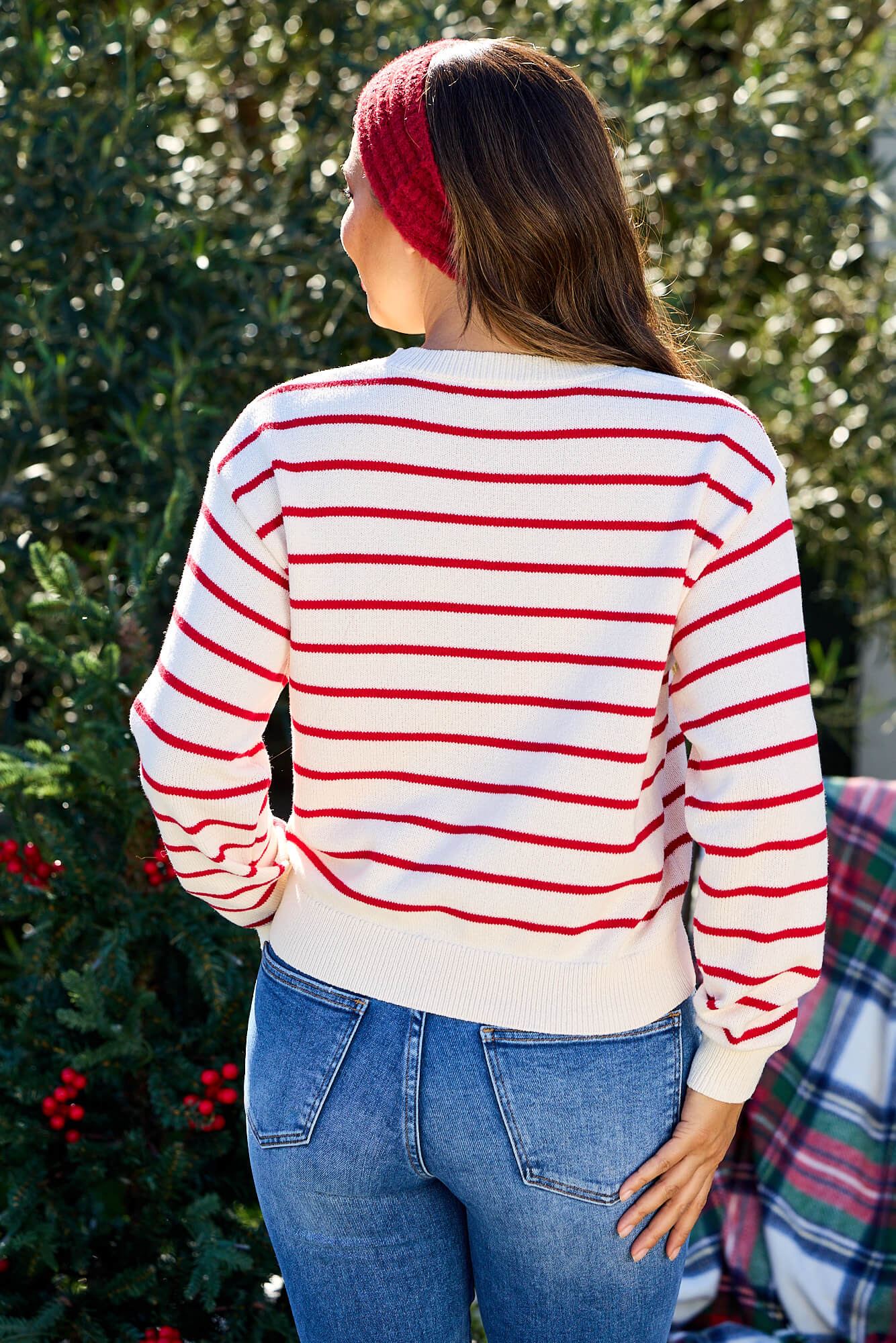 A woman outdoors faces away, wearing a Cream and Red Striped Raglan Sleeve Sweater - FINAL SALE, with blue jeans and a red headband.
