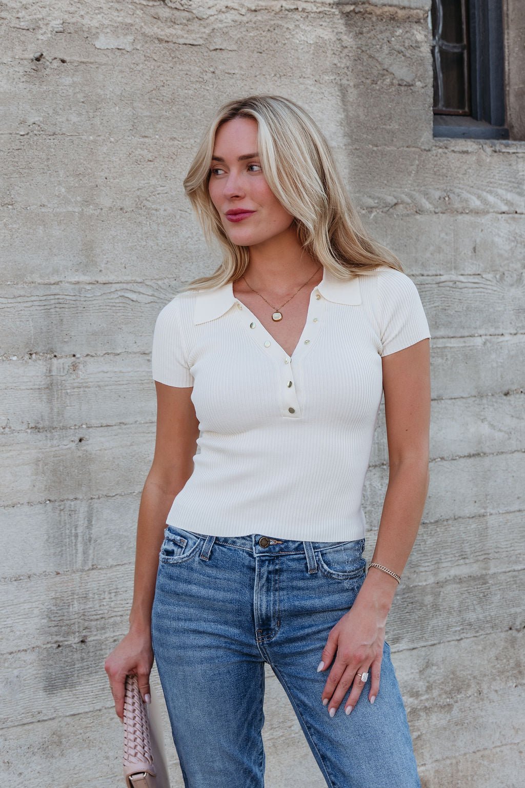 A woman in a Cream Button Up Ribbed Top - DOORBUSTER and blue jeans stands against a textured concrete wall, holding a beige clutch.