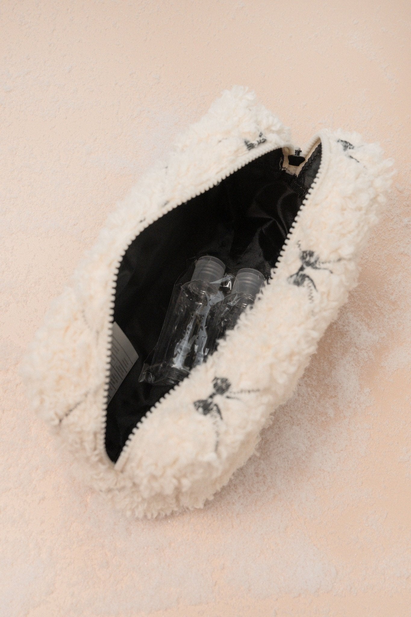 The Cream Cherie Sherpa Cosmetic Pouch Bag in white with black lining, shown partially open with two clear bottles inside.