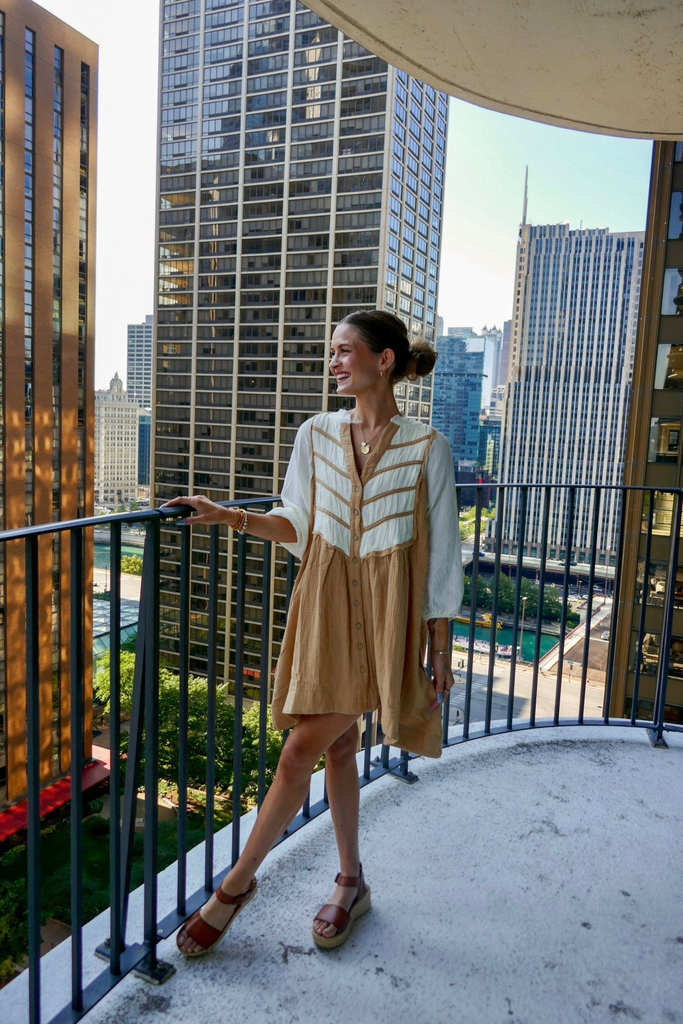 Wearing a Cream Color Block Tunic Dress, a woman smiles on a city balcony with tall buildings and trees in the background.