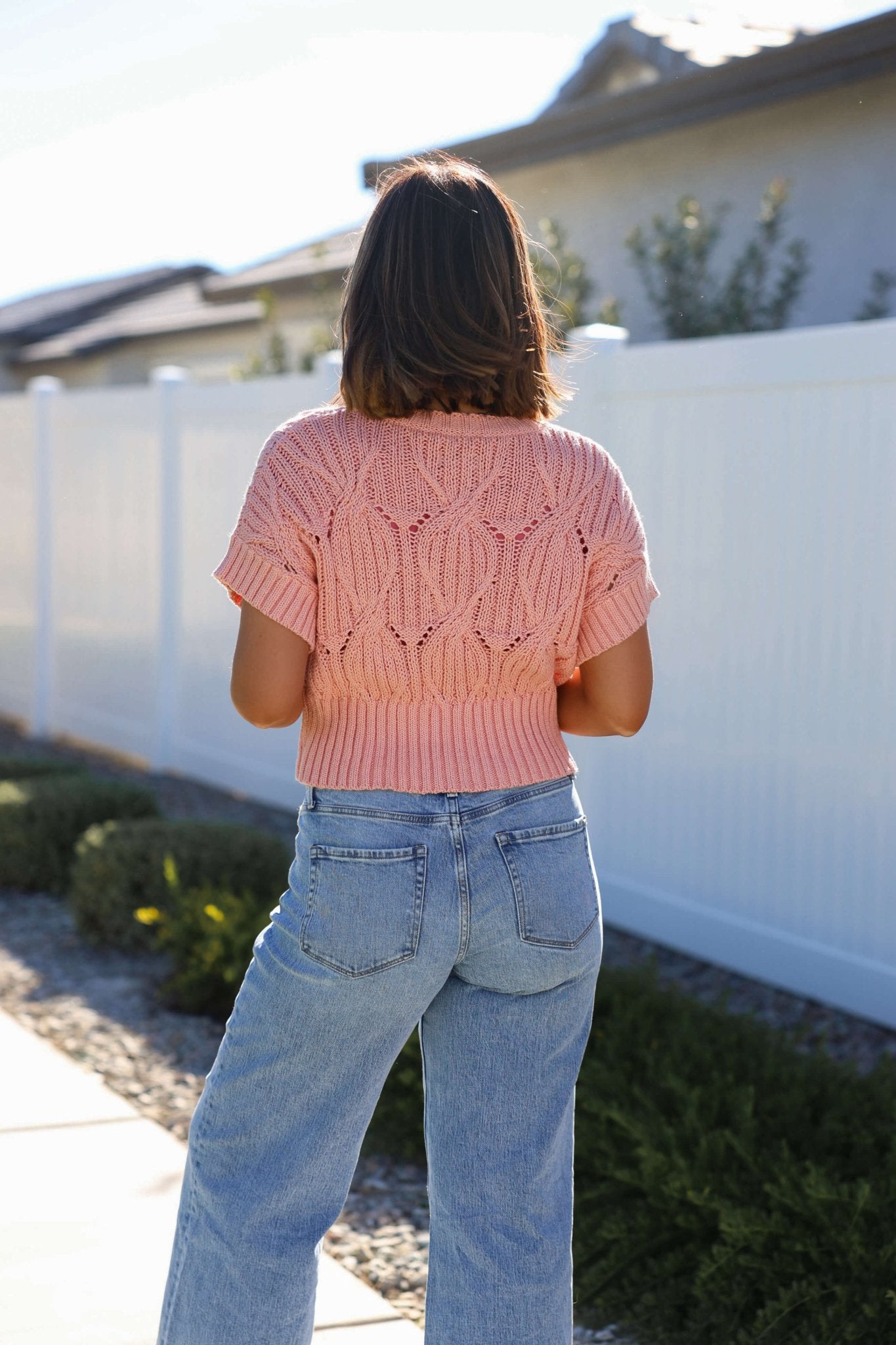 A woman in a Crochet Detail Short Sleeve Sweater - Blush and blue jeans stands outdoors by a white fence with greenery behind her.