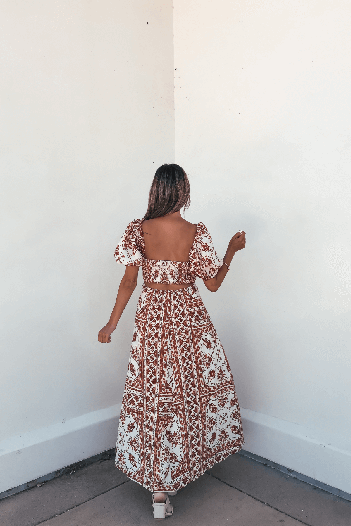 Woman with long hair in the Cynthia Multi Rust Print Midi Dress stands with her back to the camera, facing a white corner.
