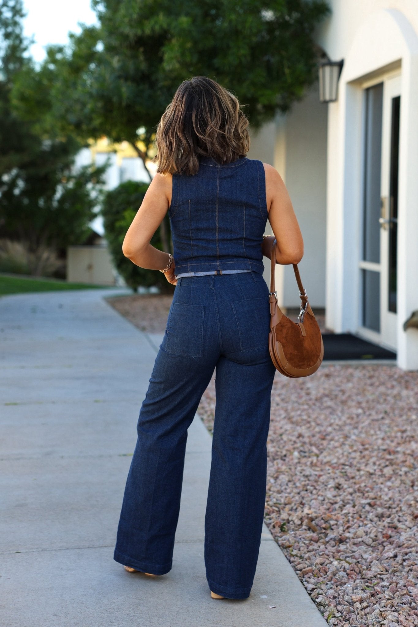 A woman in Dark Denim Front Seam Wide Leg Jeans holds a brown handbag, standing on a sidewalk facing away.