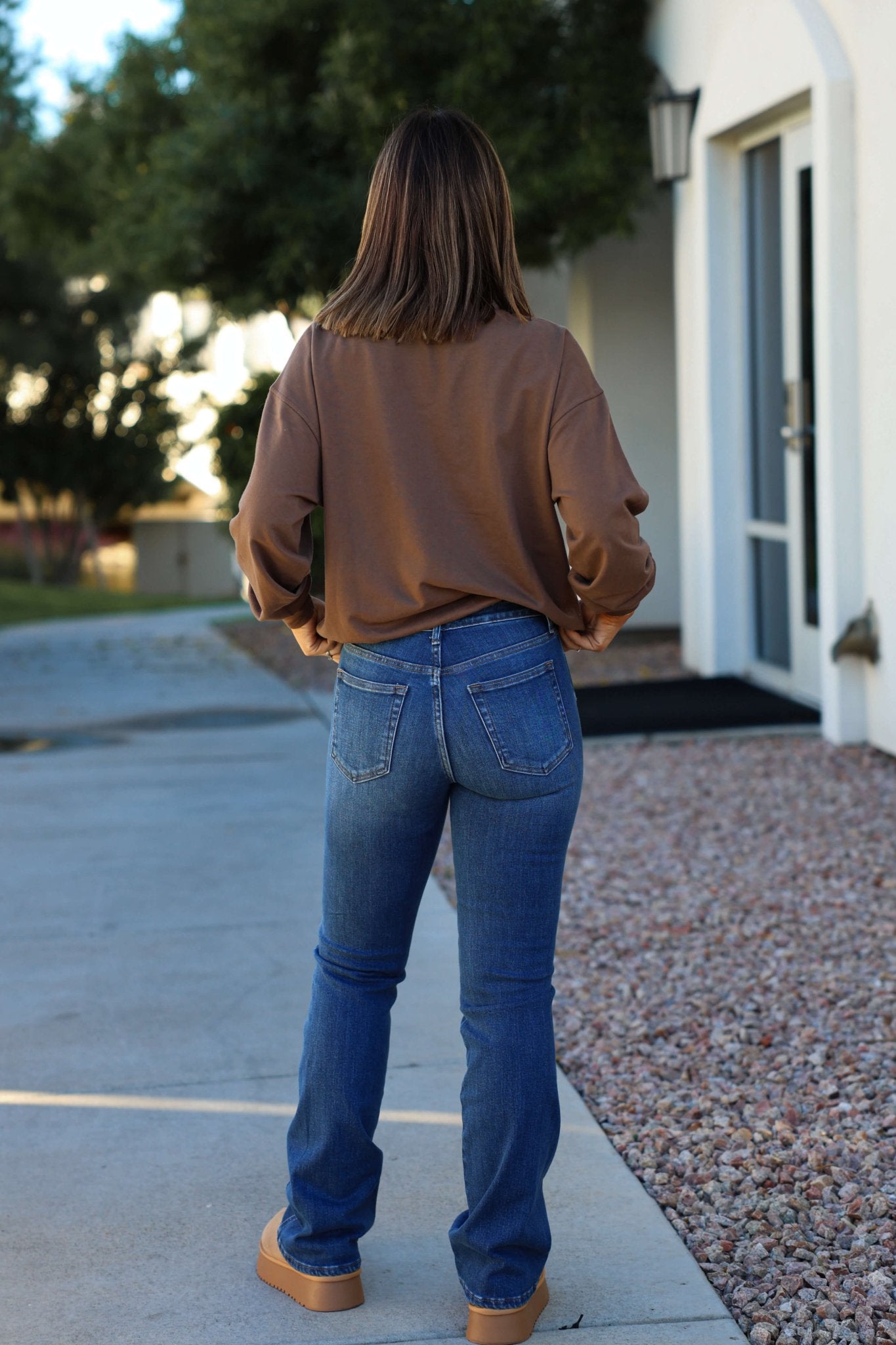A person with straight brown hair in a brown top and Dark Wash Mid Rise Bootcut Jeans stands outside on a sidewalk facing away.